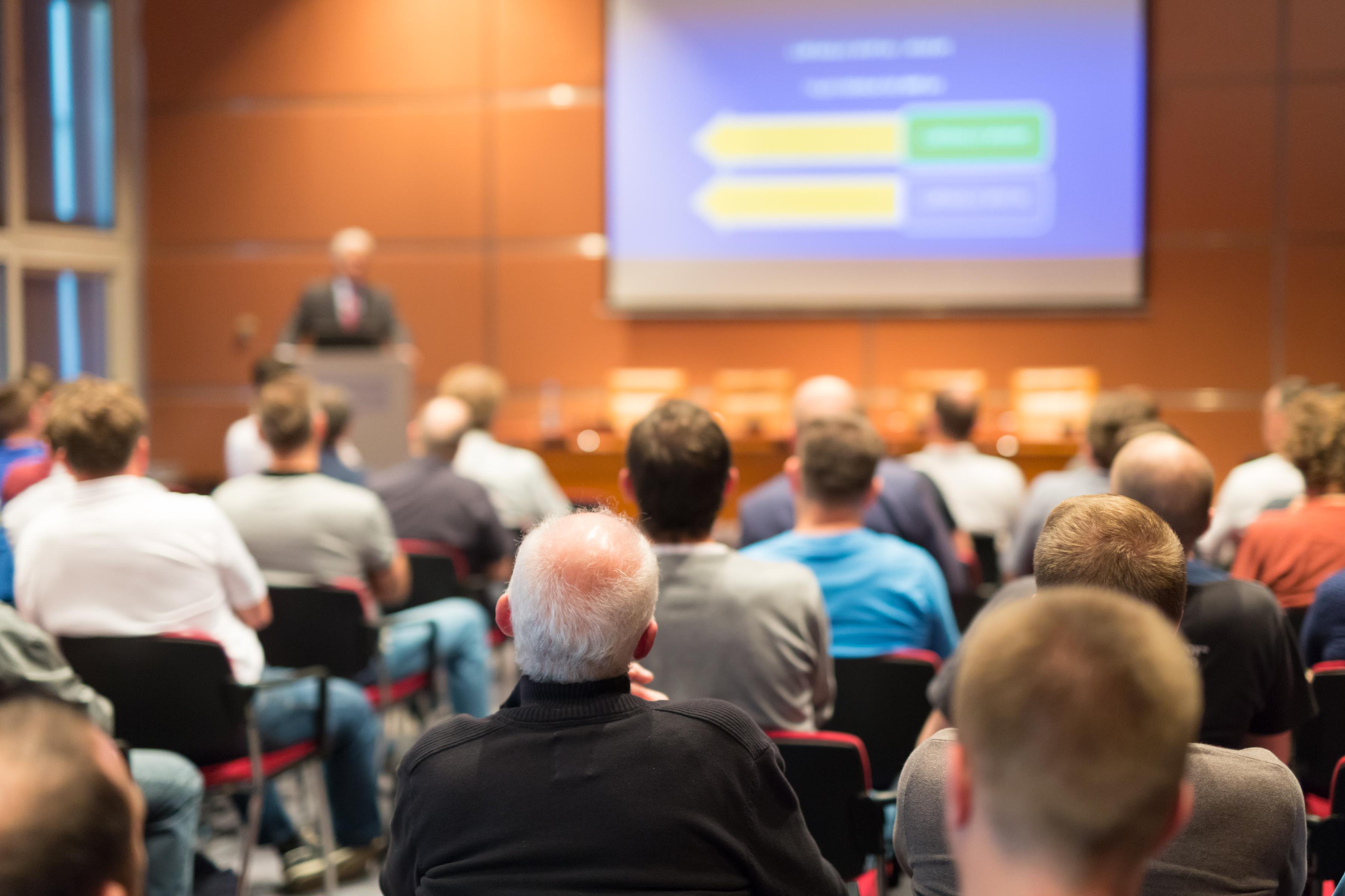 A presenter stands at the podium in front of a crowded lecture hall. Presentation slides are projected on a screen behind them.  