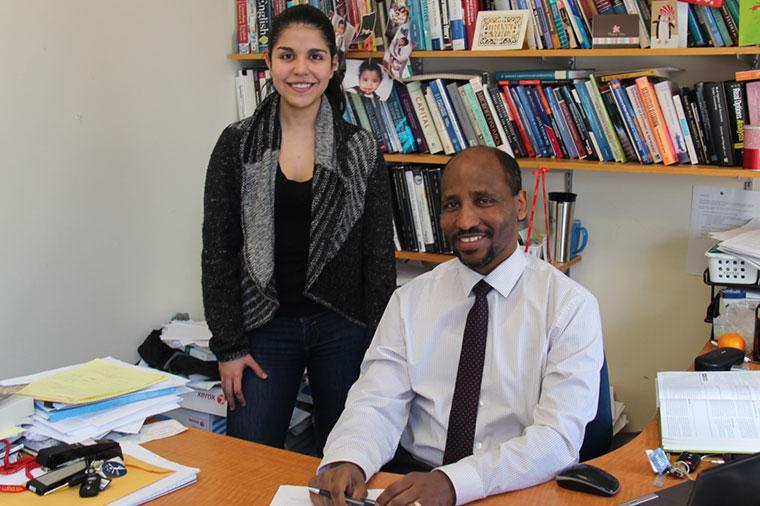 Female student stands beside male professor at desk with books behind them