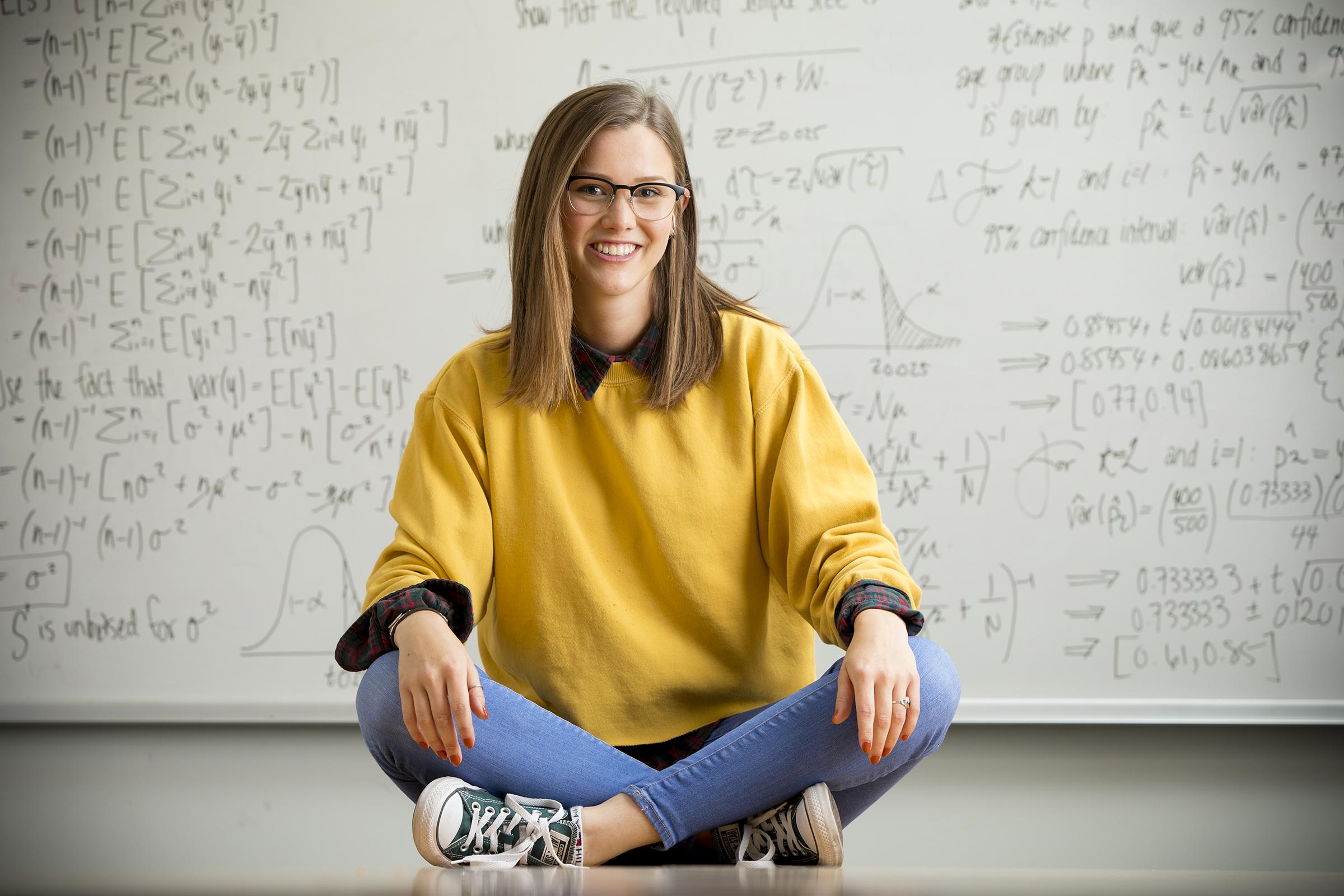 A woman sits in front a white board of complex math equations