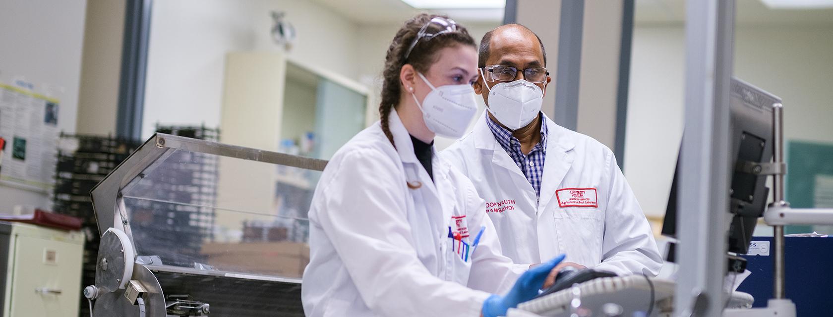 Lab technicians in masks and lab coat working closely at a machine.