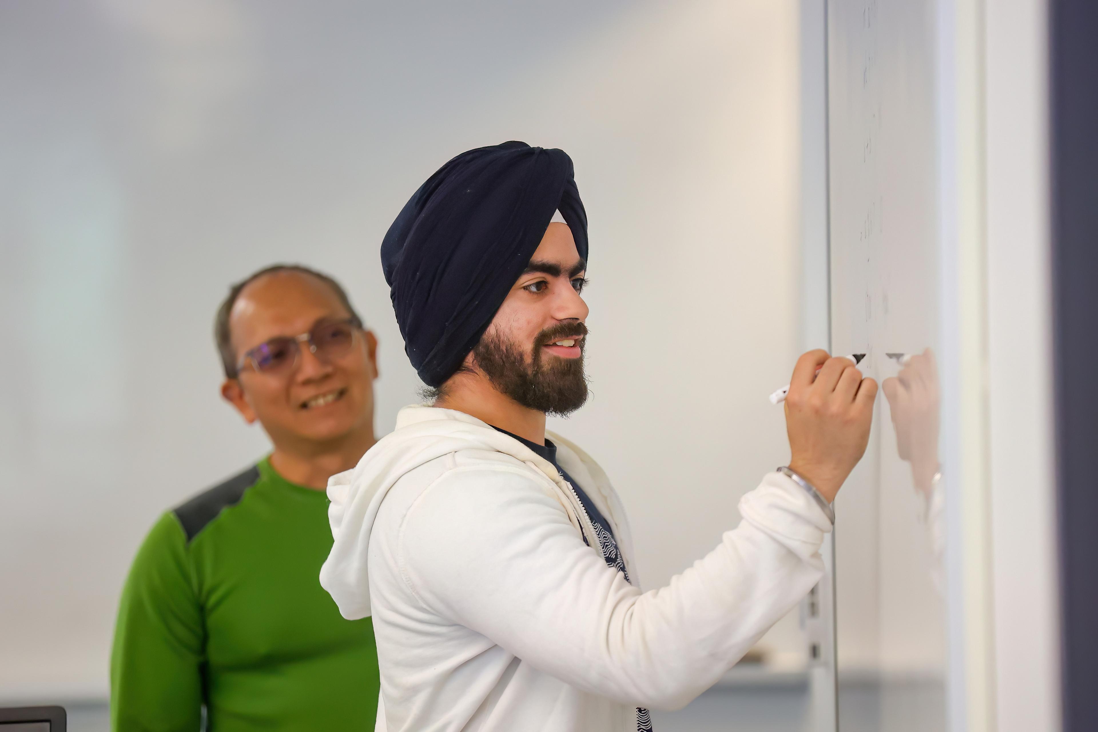 A computer science student writing on a whiteboard as professor watches smiling.