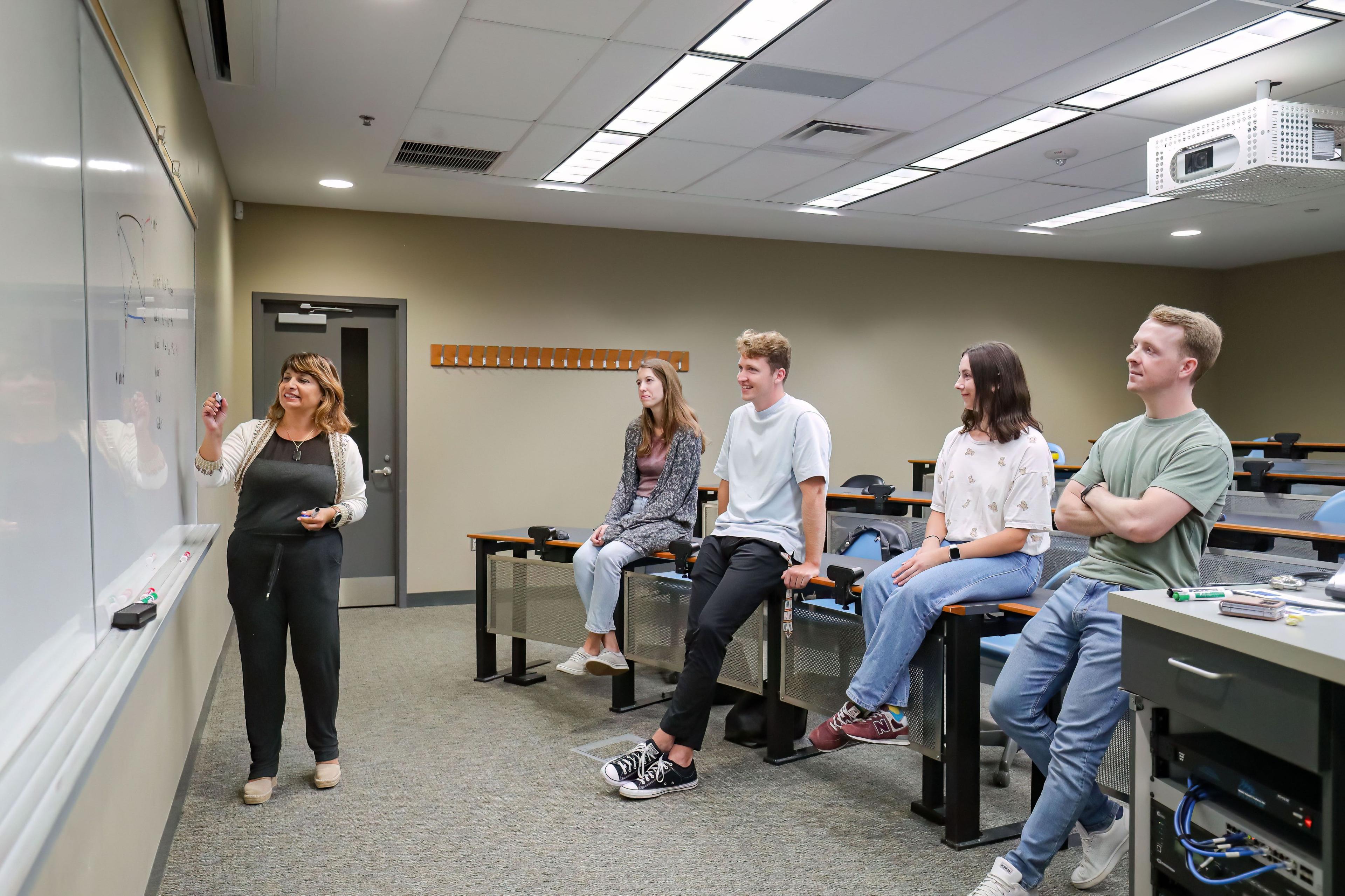 4 Students sitting on desk in a classroom watching professor as she writes on the whiteboard.