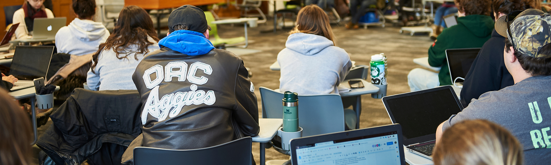 Showing the backs of a group of students in a classroom, where one student is wearing an "Aggies" jacket.