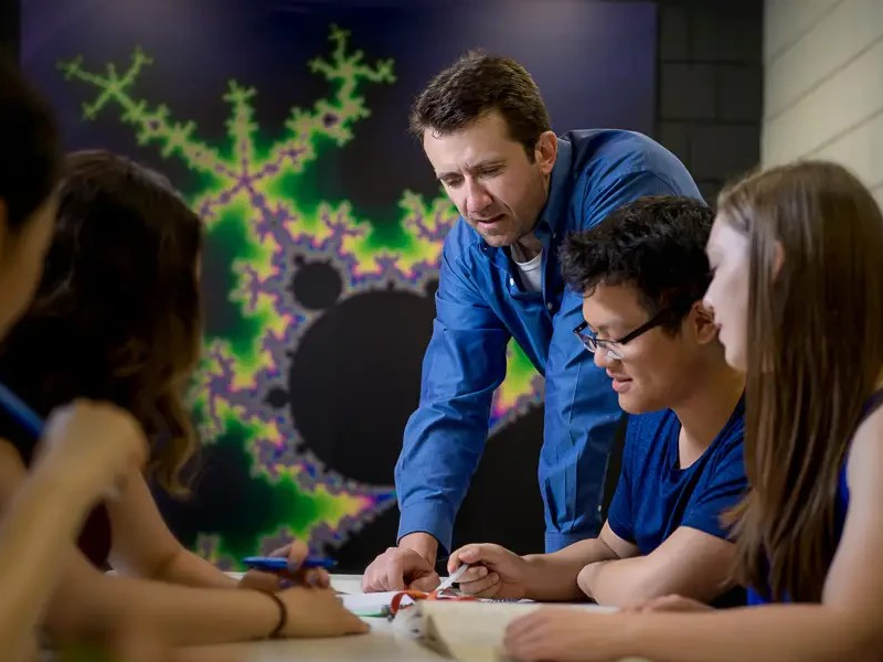 Group of students gathered around a table, one student pointing at a paper while explaining a concept.