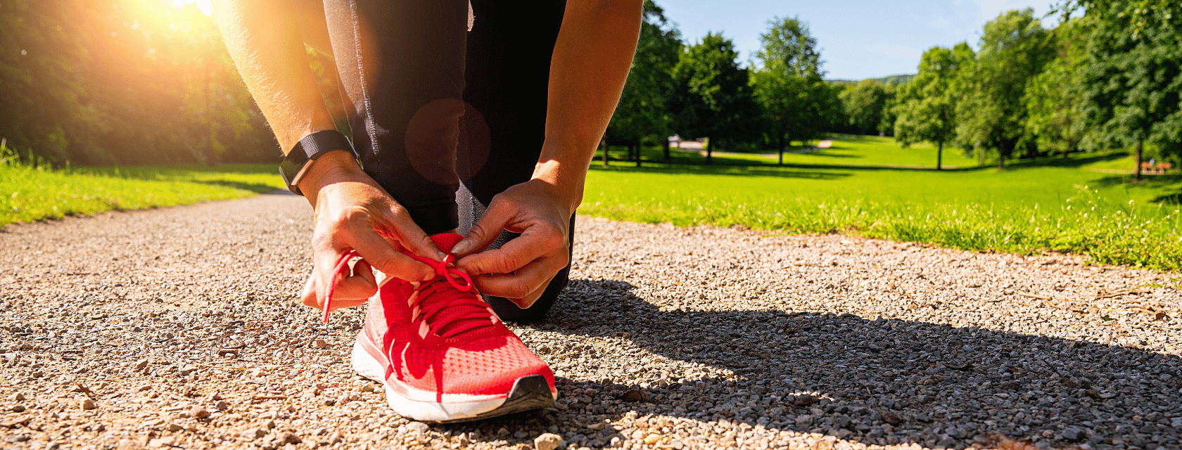  A person tying the laces of a bright pink running shoe on a gravel path in a park surrounded by green trees and grass under a sunny sky.
