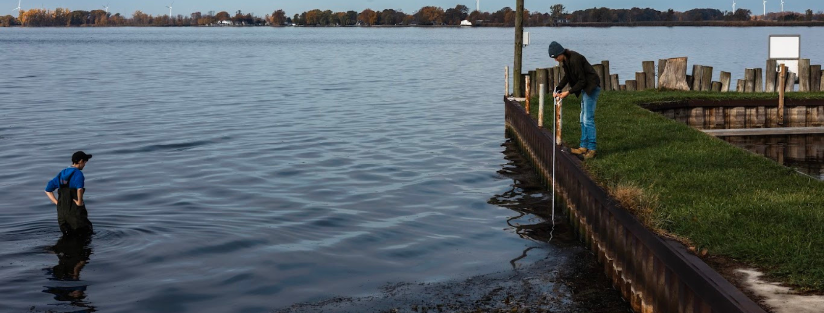 An environmental technician student standing in shallow water with waders on observing another student standing on land, lowering a secchi disc into water to measure turbidity.