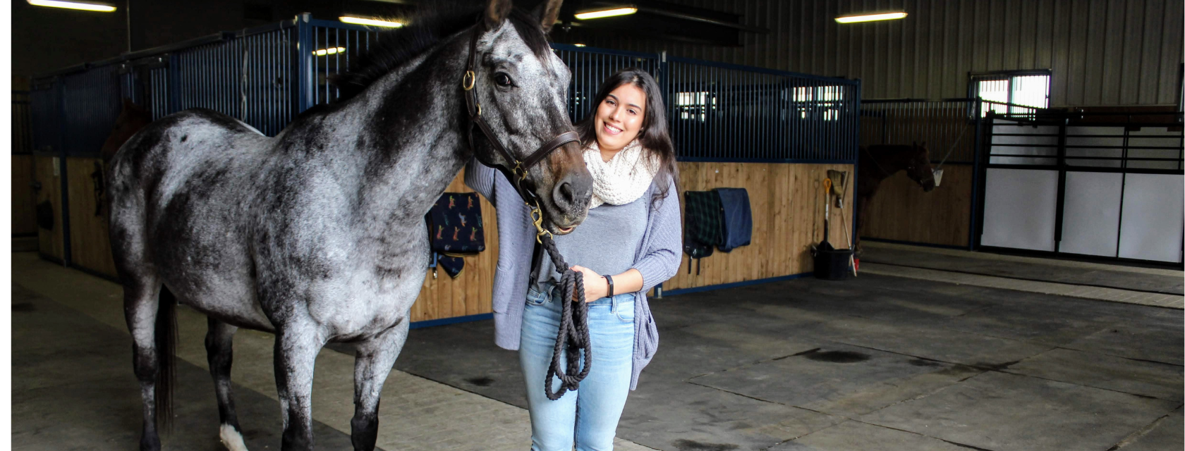 Equine care and management student standing alongside a horse while smiling. The background features horse stalls. 