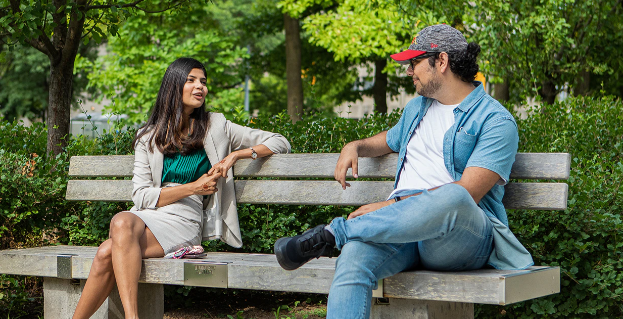 Two University of Guelph Students sitting on a bench4 at a distance from each other, chatting.
