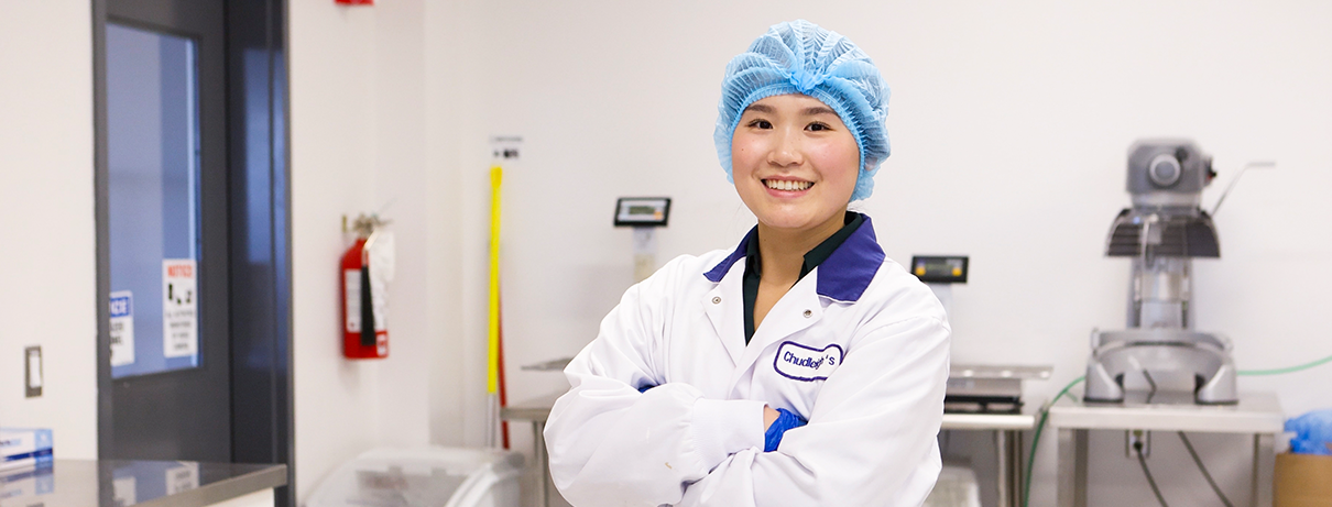 aleana standing in a food lab, arms folded, wearing a lab coat and hair net