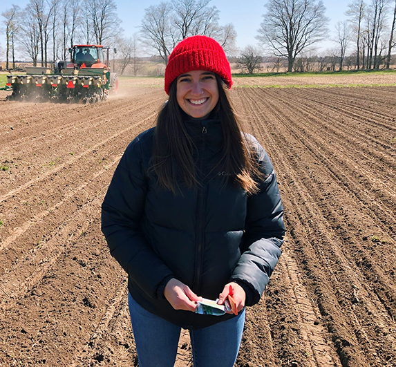 mikayla standing in a field with a tractor behind her, plowing