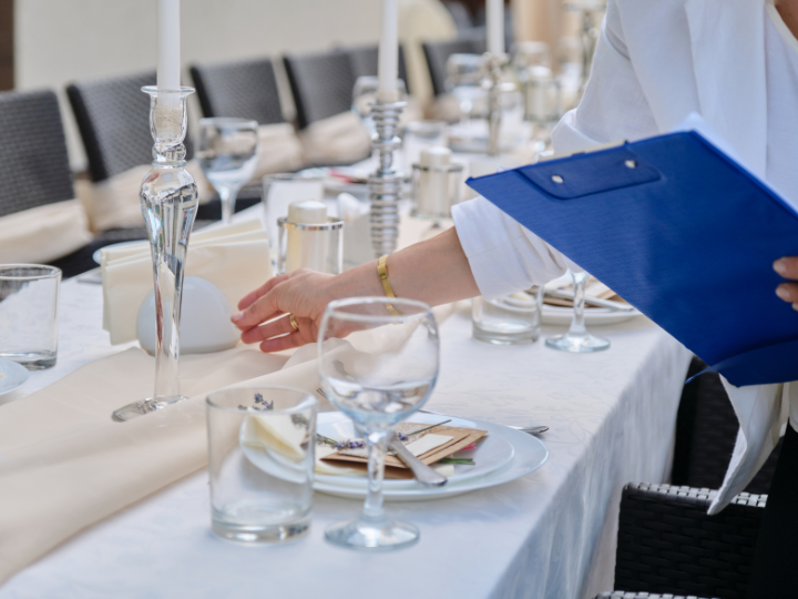 a person holding a clipboard and adjusting items on a table set for an event.