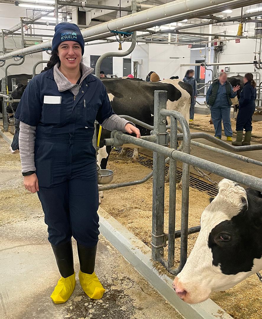 Individual in overalls and a hat standing in a dairy barn, smiling at the camera, with a cow nearby; other people in background.