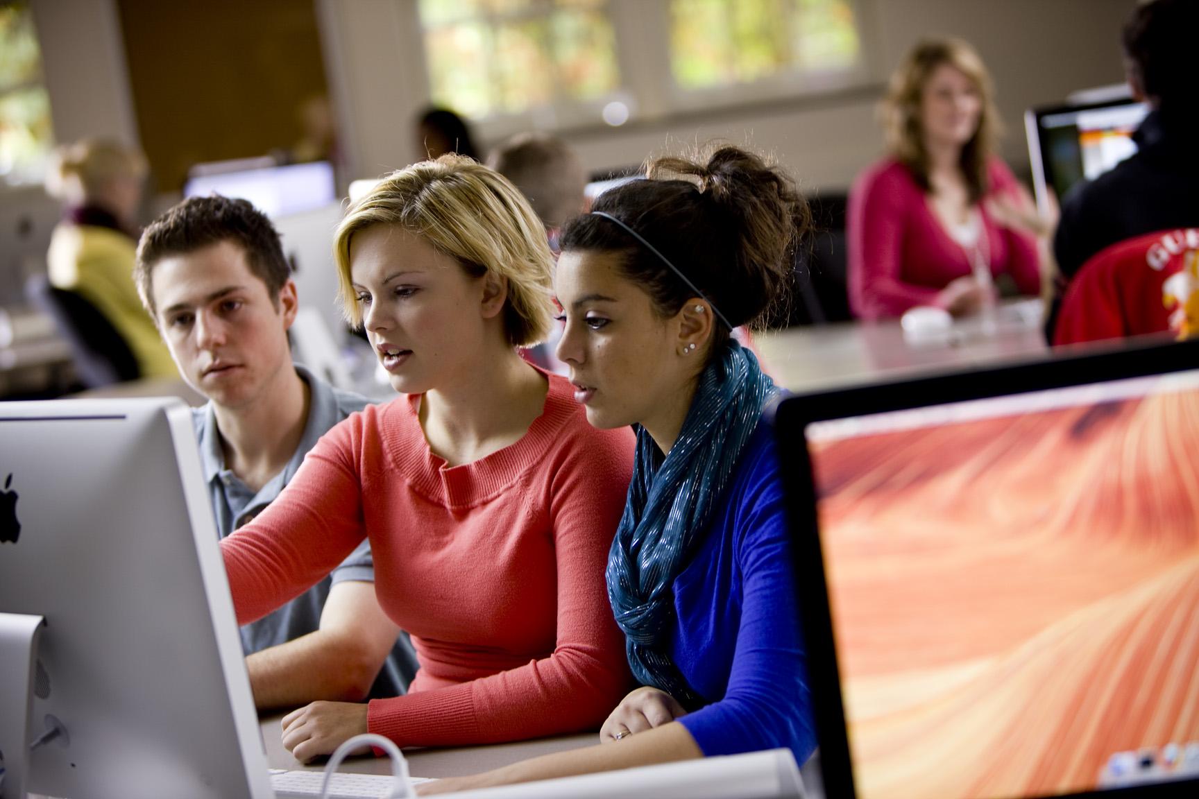 Three students sitting together at a computer, looking at screen, with other students at tables in the background.