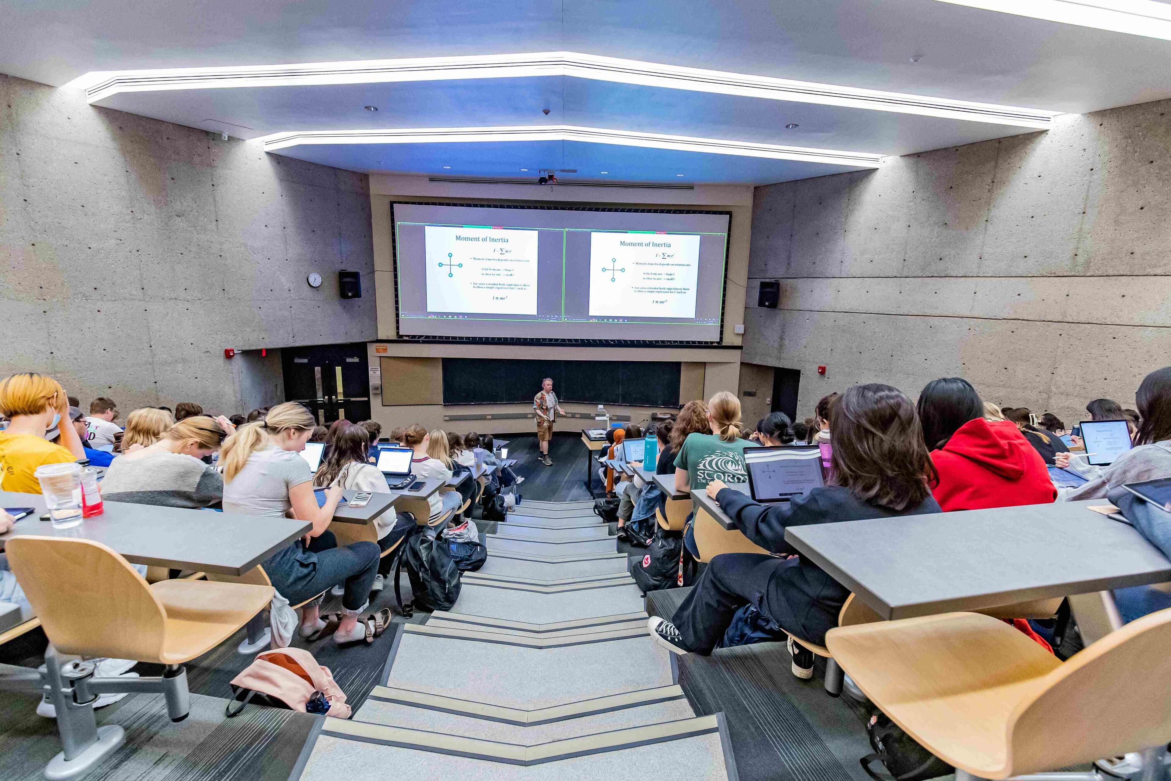 A view of a lecture hall from the back of the room facing backs of students, a prof standing at the front and a slideshow being projected.