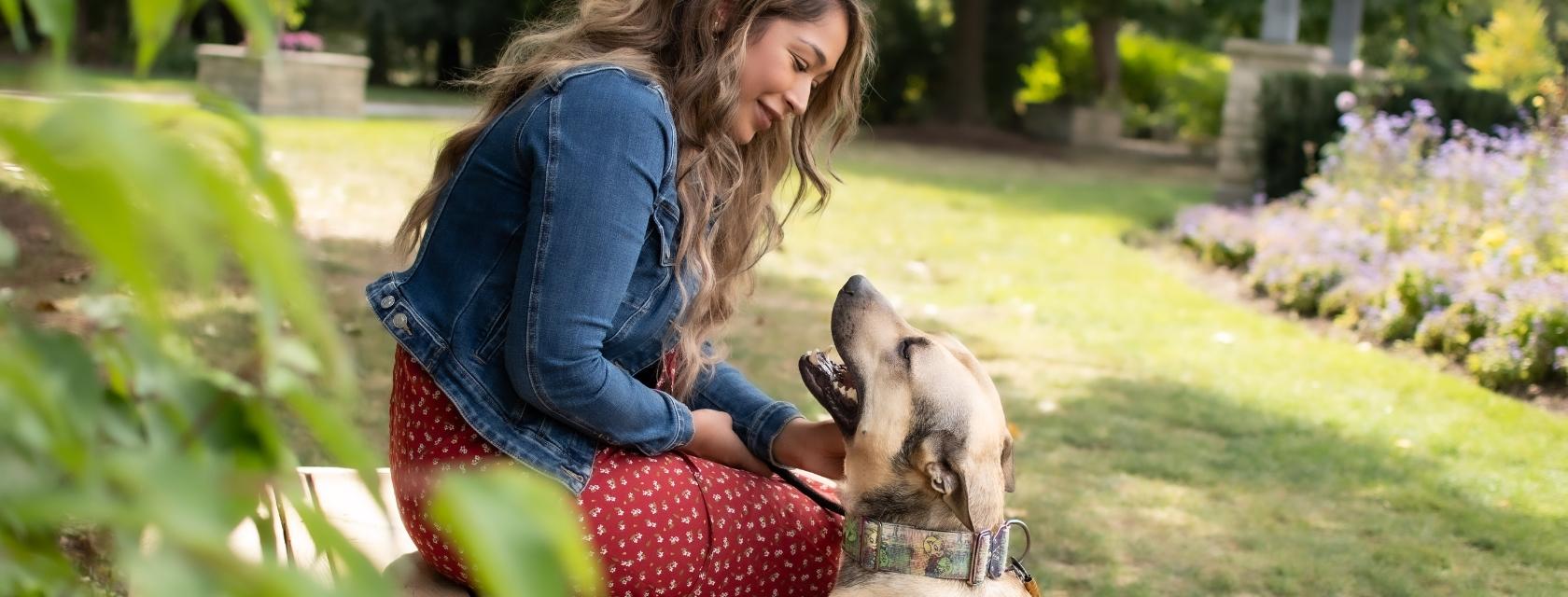 A woman wearing a jean jacket and a red dress sitting on a bench and petting a black and tan dog