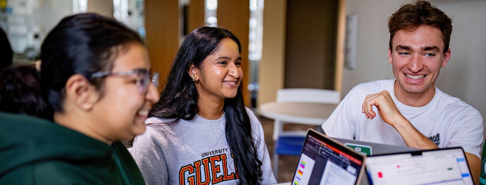 U of G students sit smiling together at a table with their laptops.