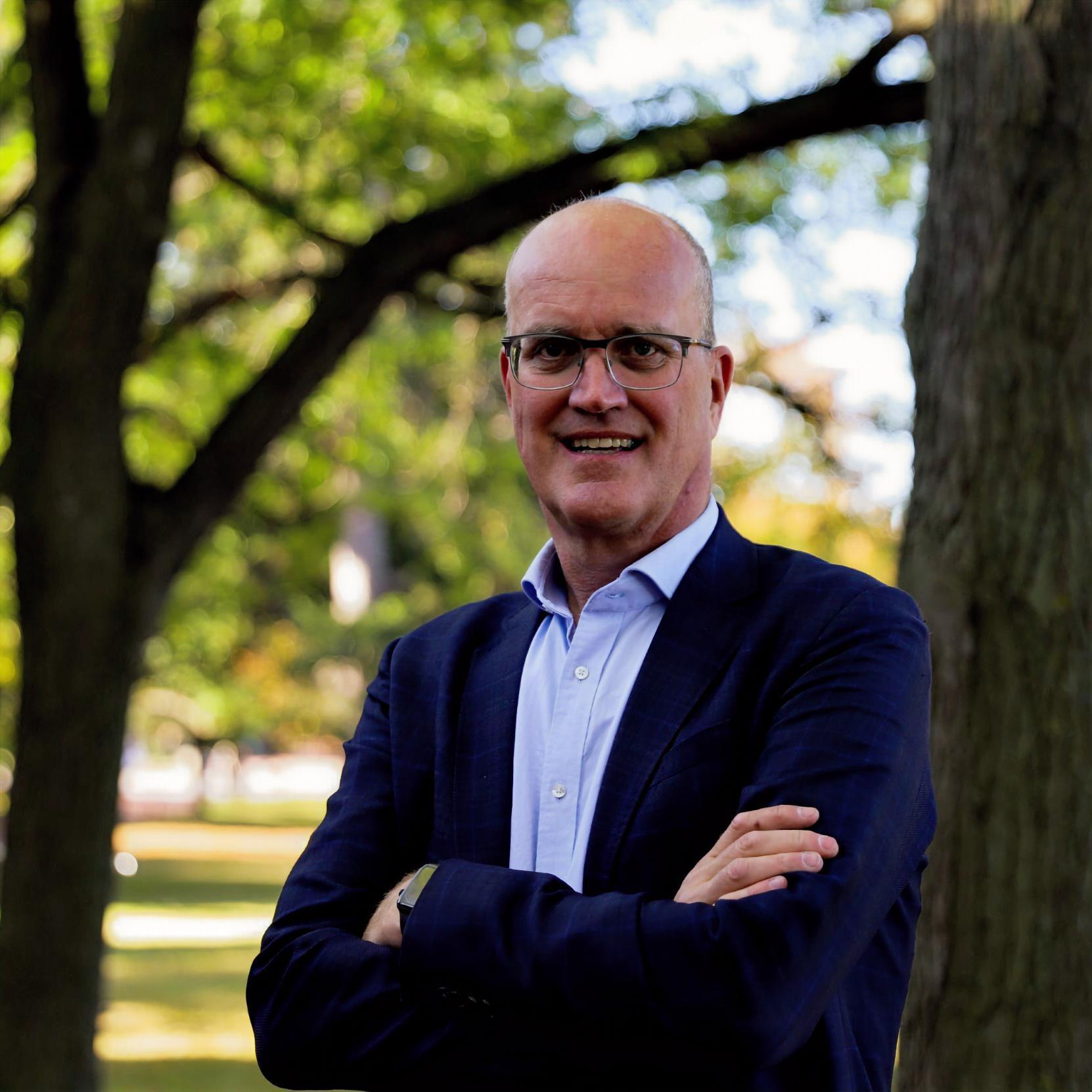 von Massow is smiling, arms crossed, in front a tree. He wears glasses, a collared shirt and a cardigan