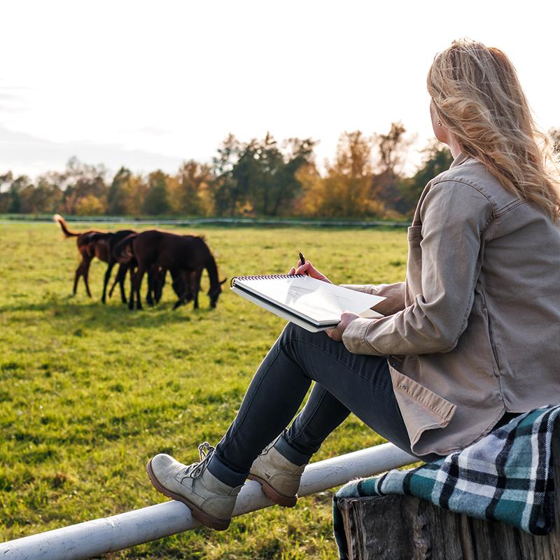 Young woman with a notepad watching horses from afar.