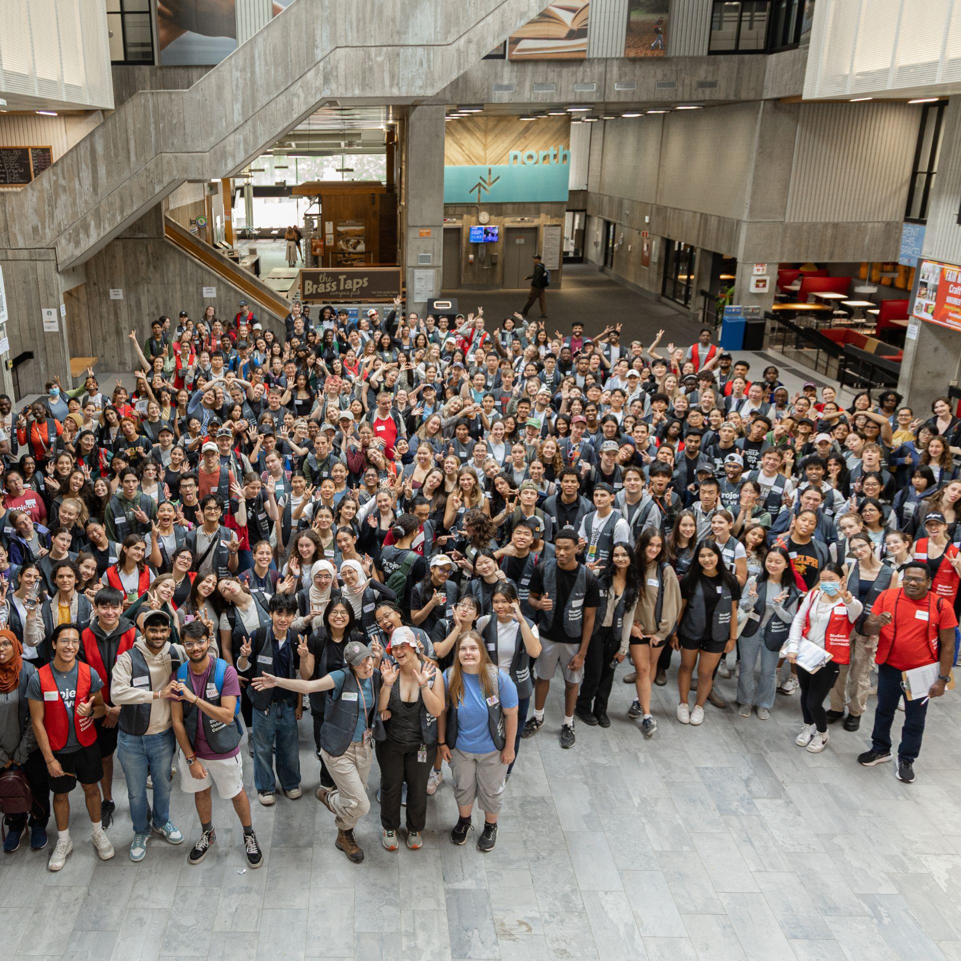 A crowd of hundred of students in the University Centre smile and wave up at a camera, taking an aerial photo.