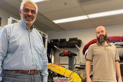 Medhat Moussa and Cole Terry standing next to a yellow robotic arm labelled "Robotics Institute" in a laboratory setting