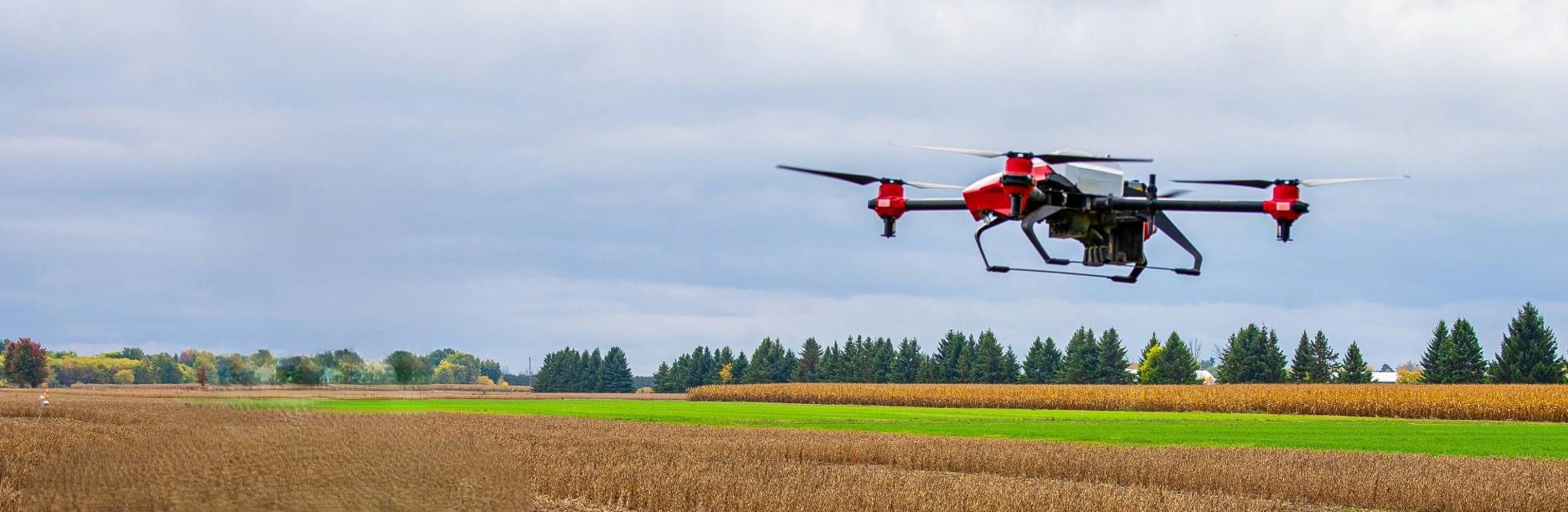 A drone with red and black features flies over expansive farmland. The landscape includes fields of crops and a line of trees under a cloudy sky.