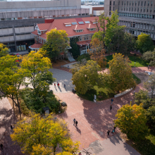 Aerial view of a university campus courtyard with brick pathways, trees with green and autumn foliage, scattered pedestrians, and surrounding academic buildings.