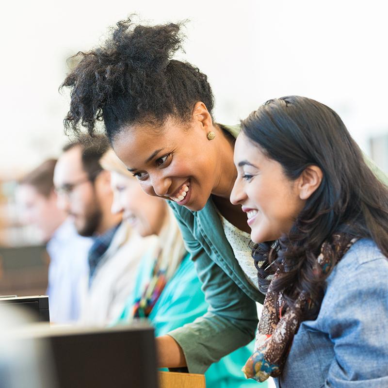 Young black woman leaning over the shoulder of a young white woman working on a computer.