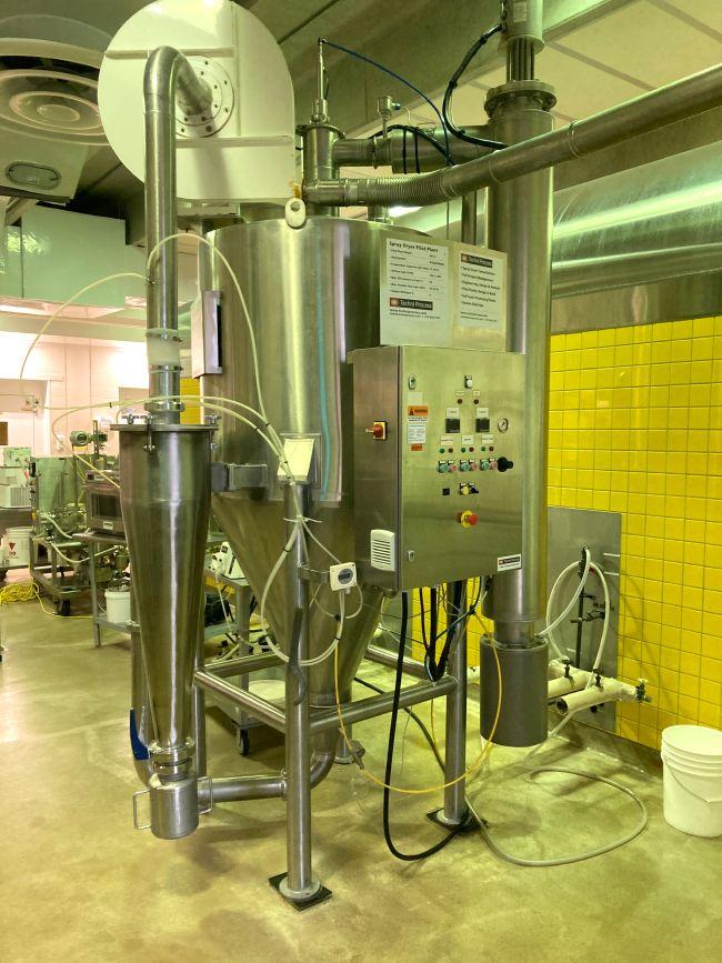 A large, shiny, cylindrical metal spray dryer with flexible piping on the outside.