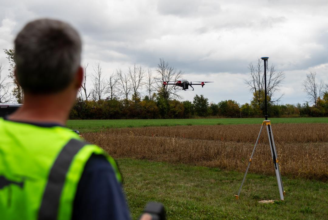 The back of Ian is visible as he flies a drone at about head height over a field