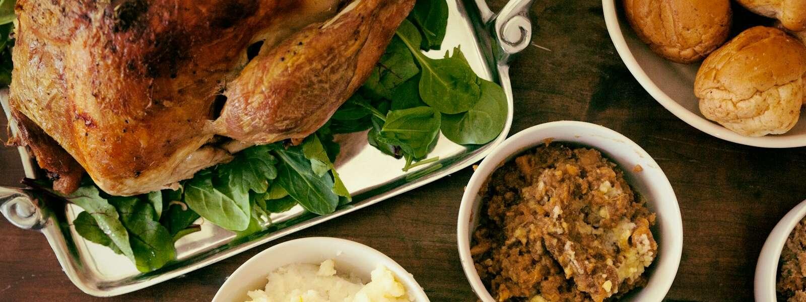 A table featuring a roasted turkey on a bed of greens, surrounded by bowls of mashed potatoes, stuffing, and a plate of rolls.
