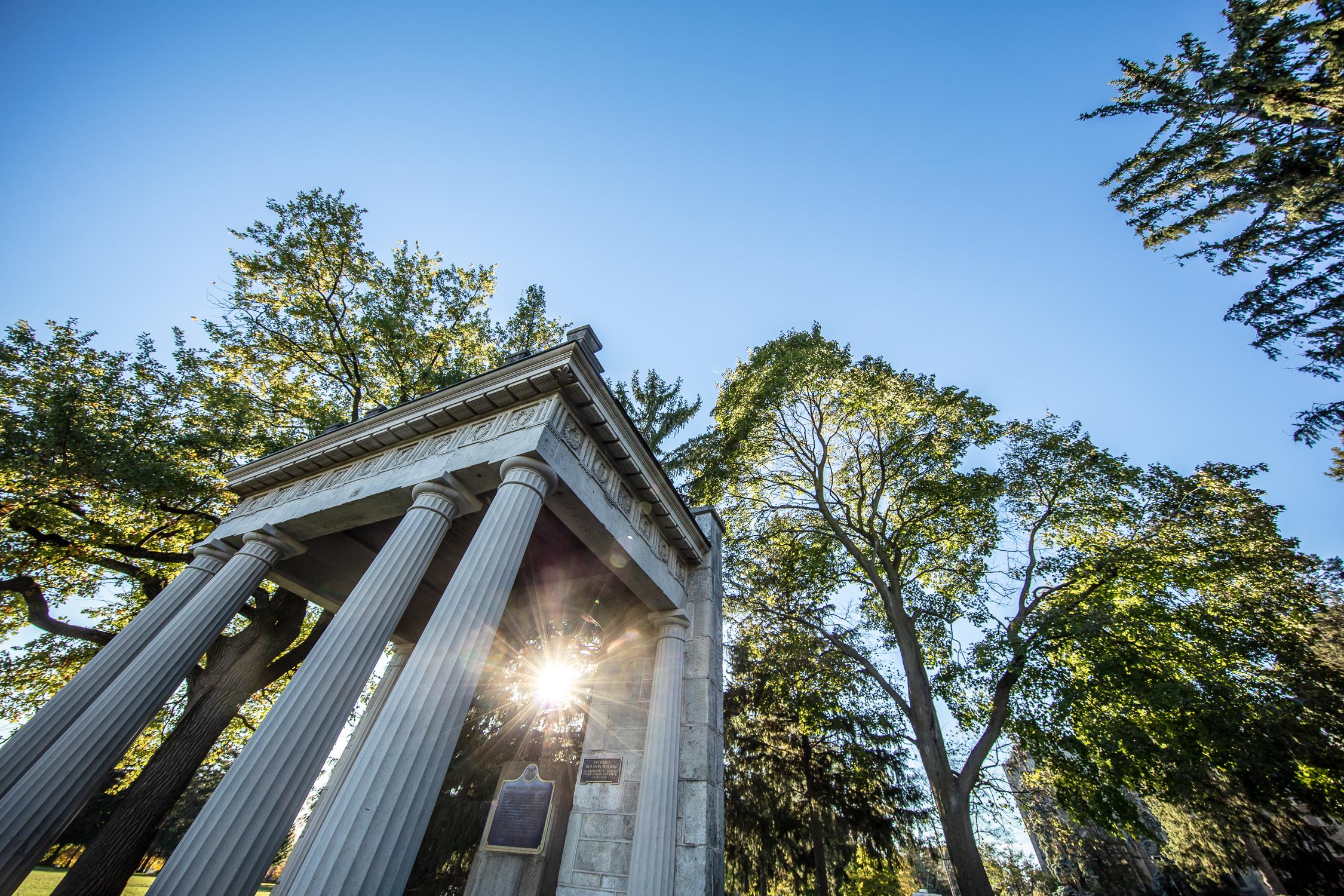 Photograph of the Portico with sun rising, peeking through the trees behind it.