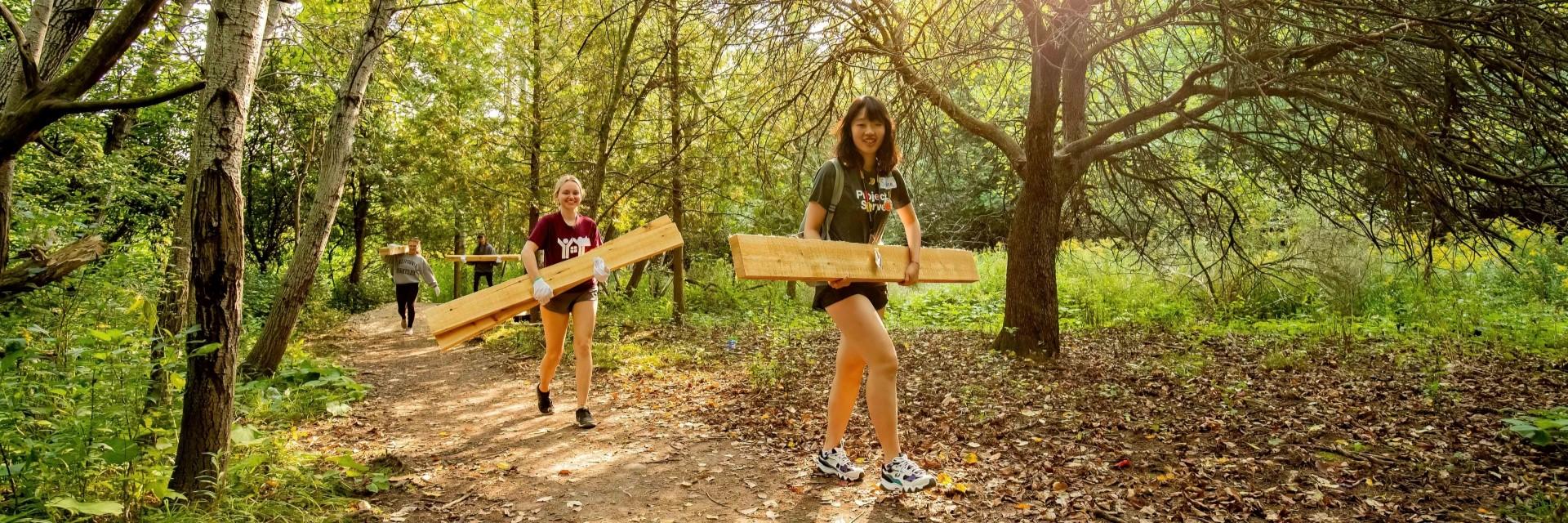 two people carrying wood planks along forested trail
