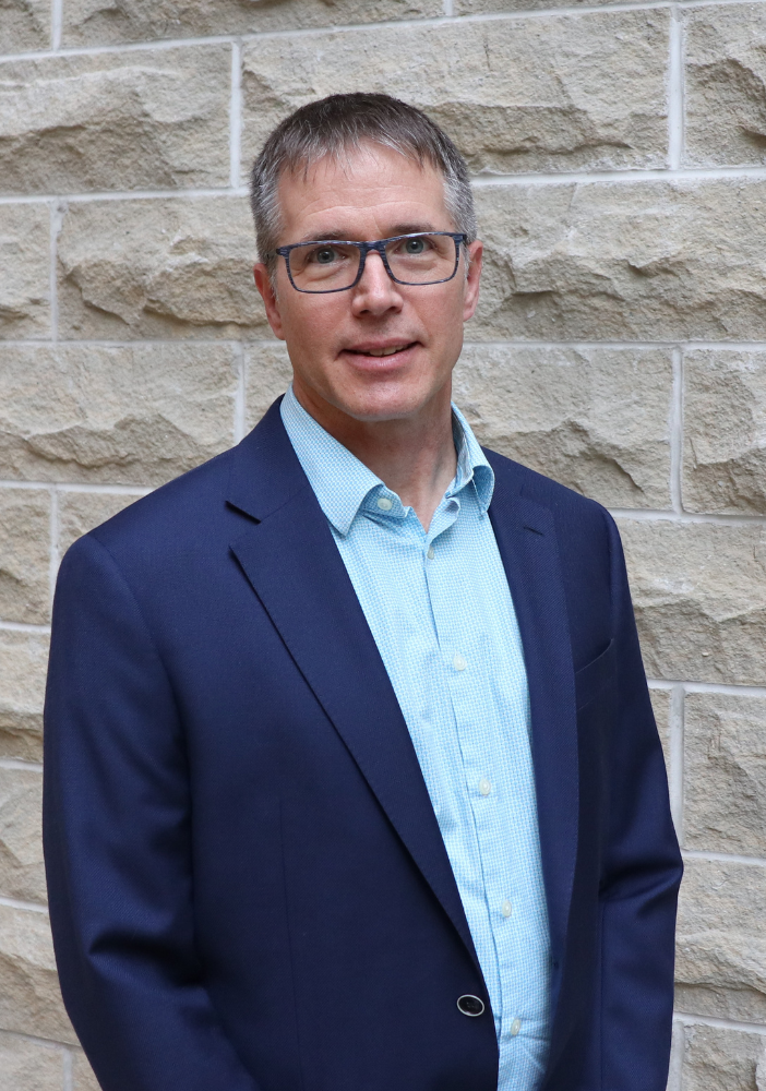 man smiling at camera in blue suite against a cream colored brick wall.