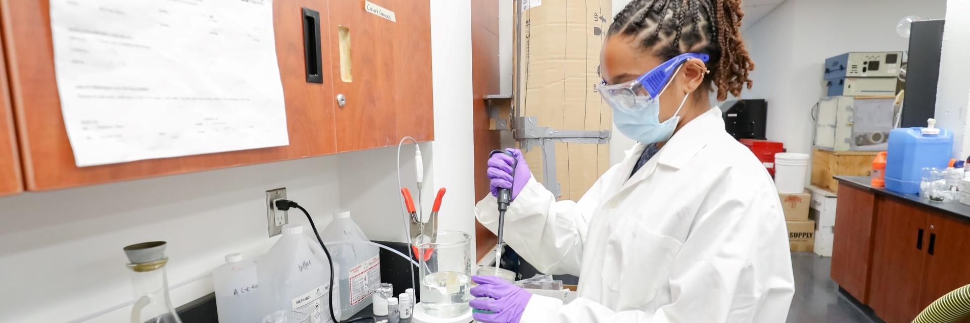 Person wearing safety goggles, medical mask, lab coat, and medical gloves uses a pipette in a lab setting.