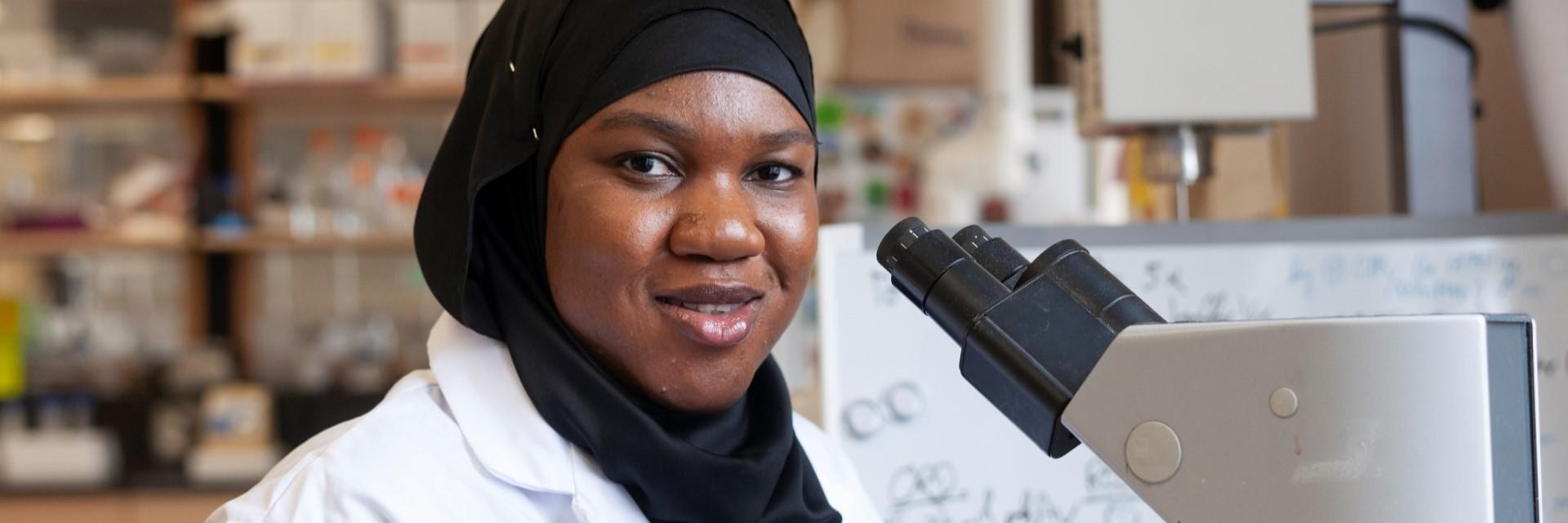 Person smiles at the camera, in front of a microscope in a lab setting.