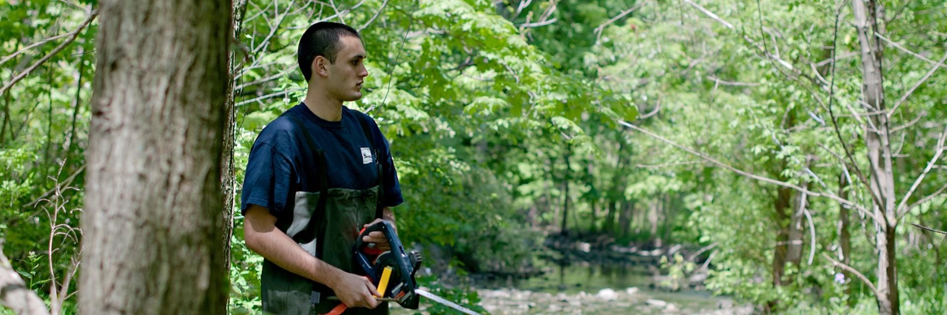 A person wearing hip waders stands outside, holding a measuring tool.