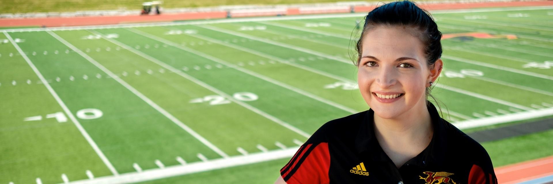 A person smiles at the camera wearing a red and black shirt, in front of a sports field.