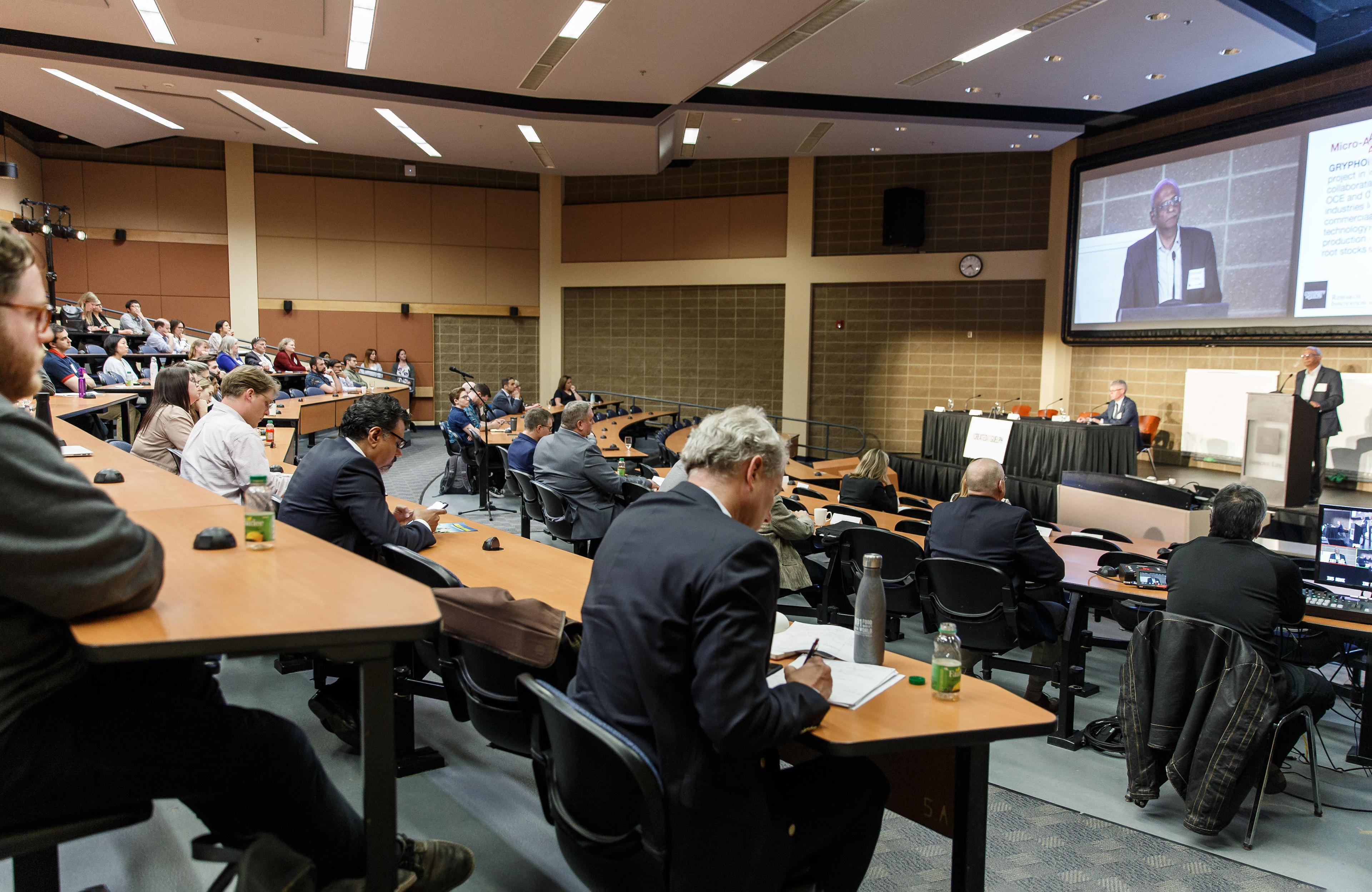 The 2019 Gryphon's LAAIR event hosted in a large lecture hall with tiered seating. Attendees, some taking notes, are focused on a presenter speaking at the podium. The speaker is also displayed on a large projection screen behind him.