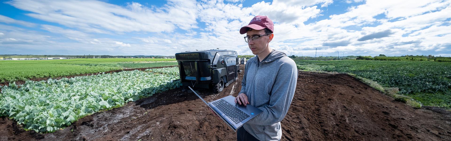 Person holding laptop while standing in a crop field with a large robotic weeder in the background.