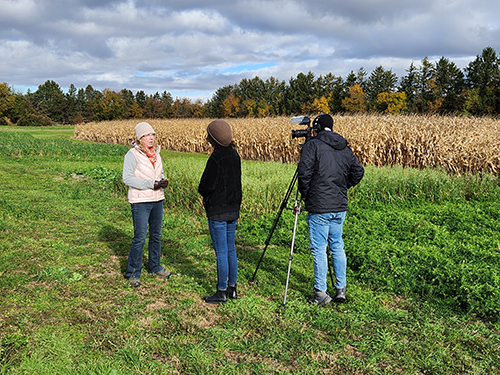 Dr. Laure Van Eerd standing in a field while being video recorded by two people.