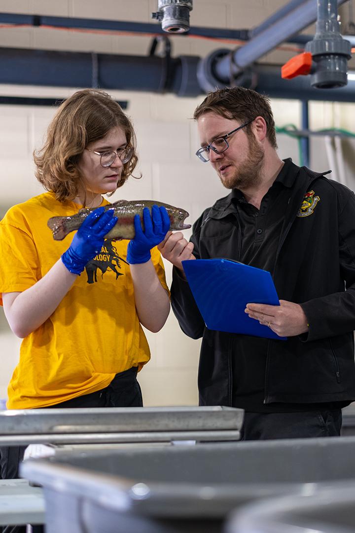Dr. David Huyben talking with student Samantha Benzer who is holding a fish, while standing in aquaculture research centre.