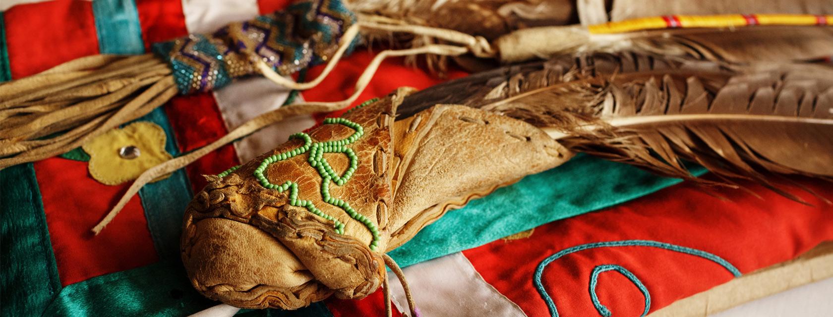 Two leather-handled feather fans. They are each adorned with colourful beading and lay on top of a red, white and turquoise fabric.