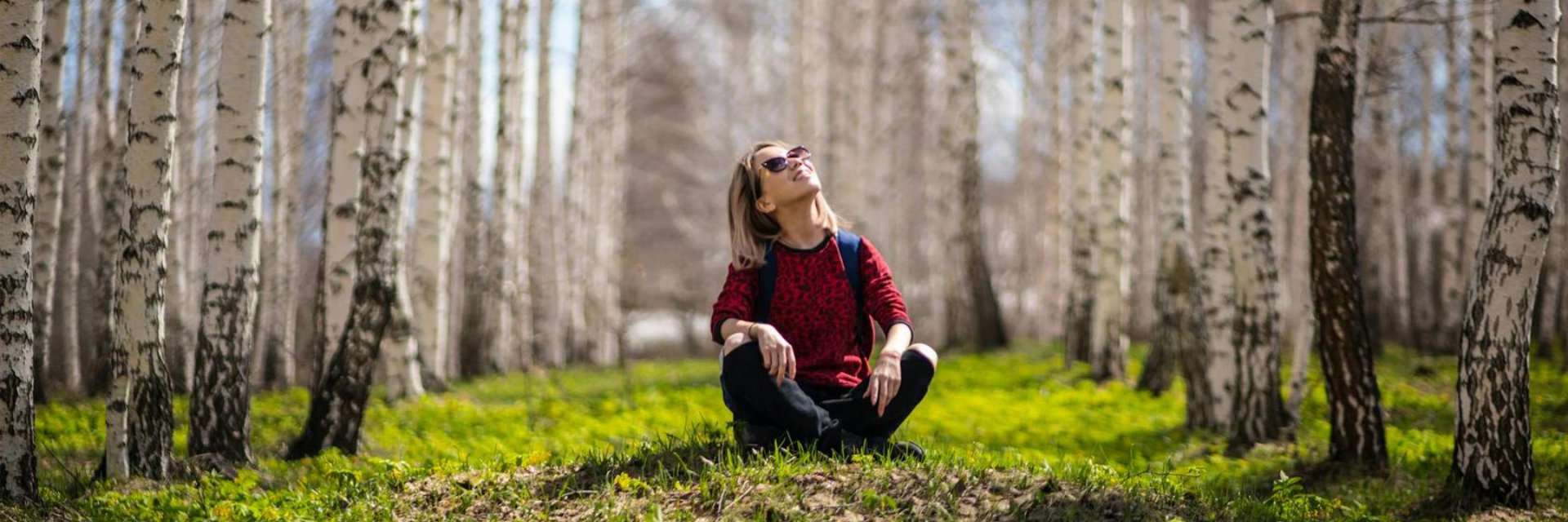 Women sitting cross legged on grass surrounded by birch trees. She is looking up at the sky