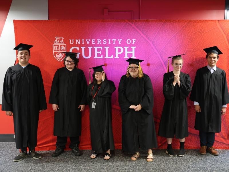 Six graduates of Project Search pose in front of a University of Guelph photo backdrop, wearing their caps and gowns.