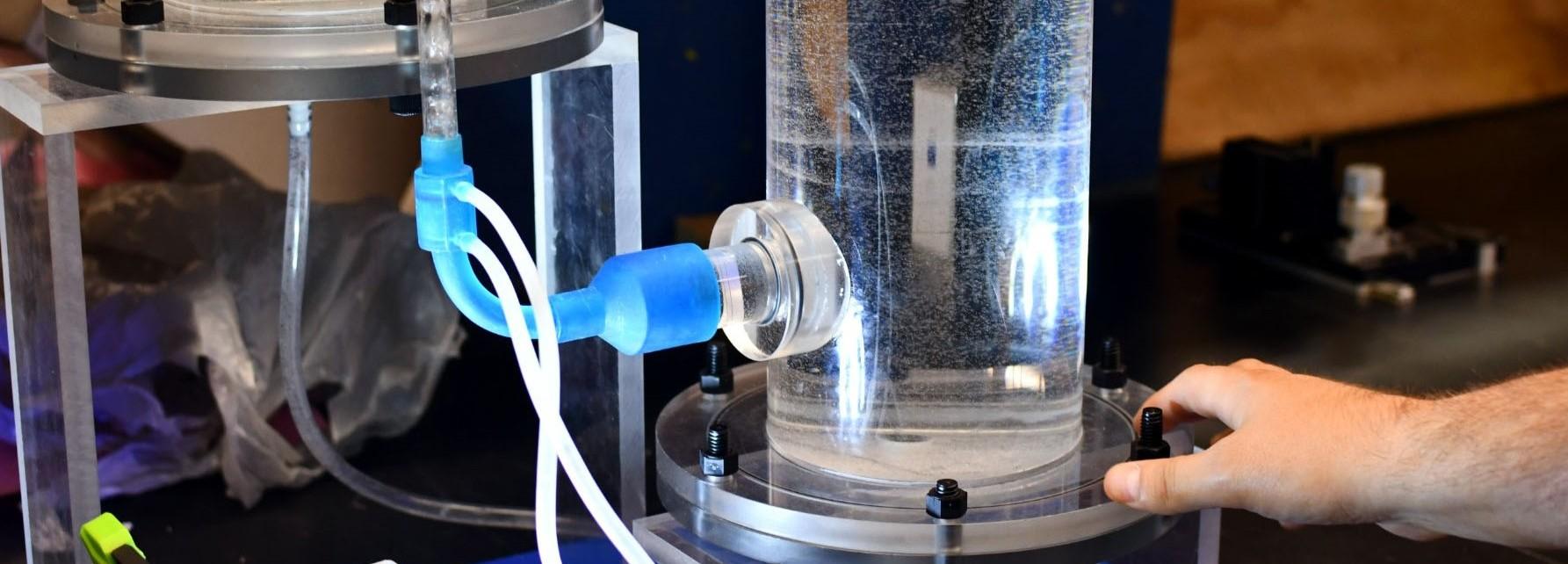 Lab setup with clear water-filled cylinder, blue tubing, and a hand adjusting the base.
