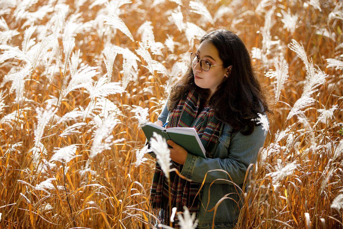 Student stands in a field of tall grass and takes hand written notes.