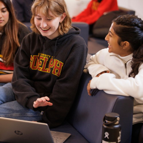 A group of students at the University of Guelph sitting together, smiling and collaborating over a laptop. The students are dressed in casual clothing, wearing university-branded sweatshirts, and engaged in a friendly conversation on campus