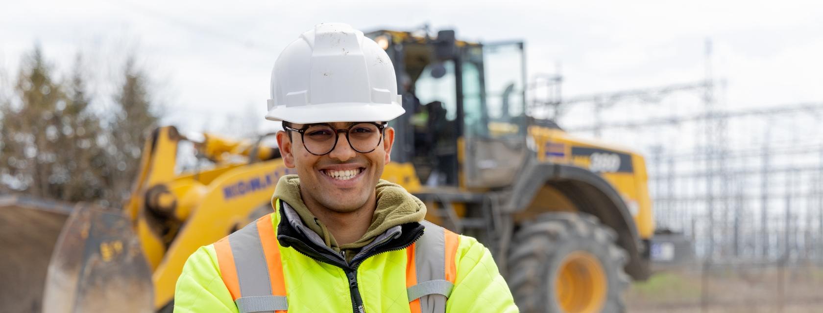 A student wears a white hard hat and safety vest. He stands in front of heavy construction machinery. Power station infrastructure is in the background.