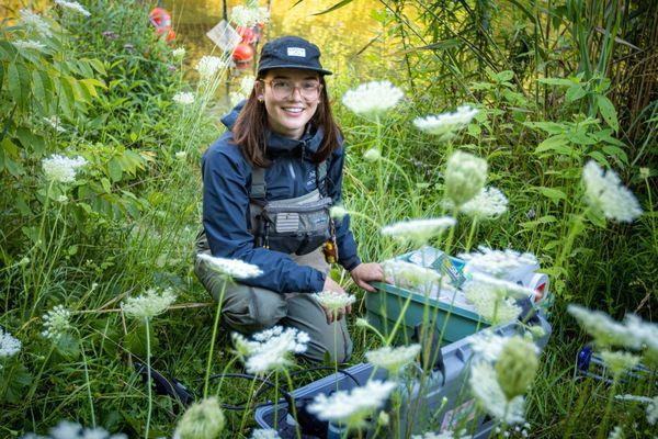 A student wears field research gear and sits among plants and environmental surveying gear.