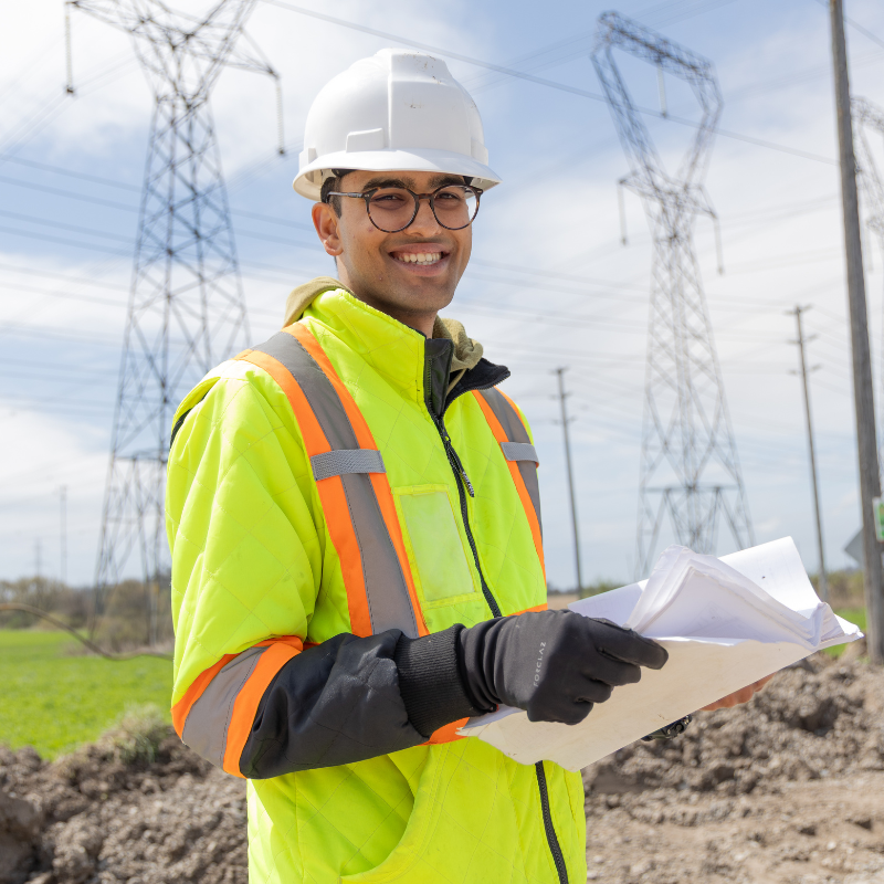 An engineering student at an outdoor job site wears a hi-vis jacket and hard hat. He holds papers, and smiles at the camera. The background shows tilled dirt from a job site, and a hydro corridor.